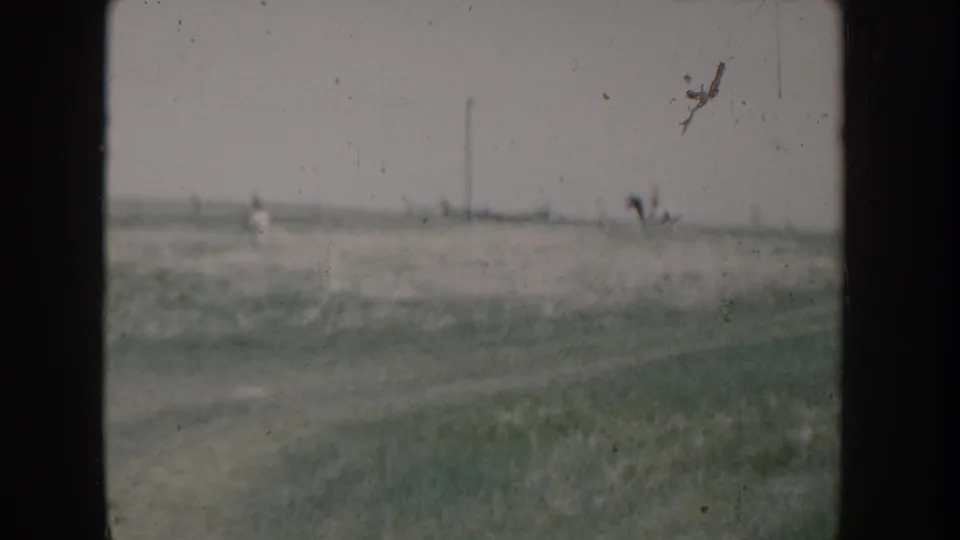 Wide 8mm home movie shot of two figures on a grassy hillside in rural Idaho, circa 1955, with a utility pole and distant fence line visible.