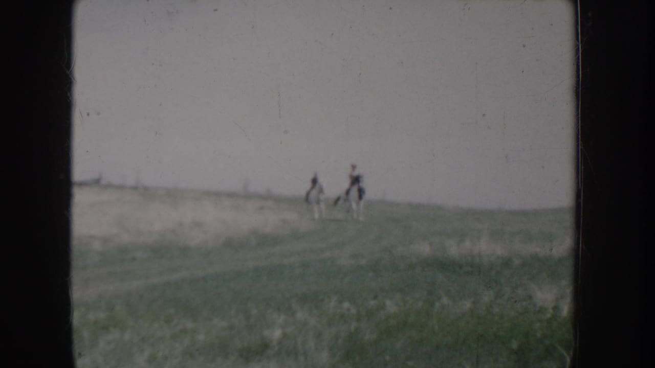 Two figures on horseback silhouetted on a hillside ridgeline against an overcast sky; wide open grassy terrain.