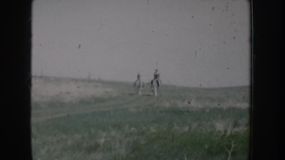 Two riders on horseback crossing an open grassy field on 8mm home movie film, Idaho circa 1955; one horse appears lighter in color than the other.