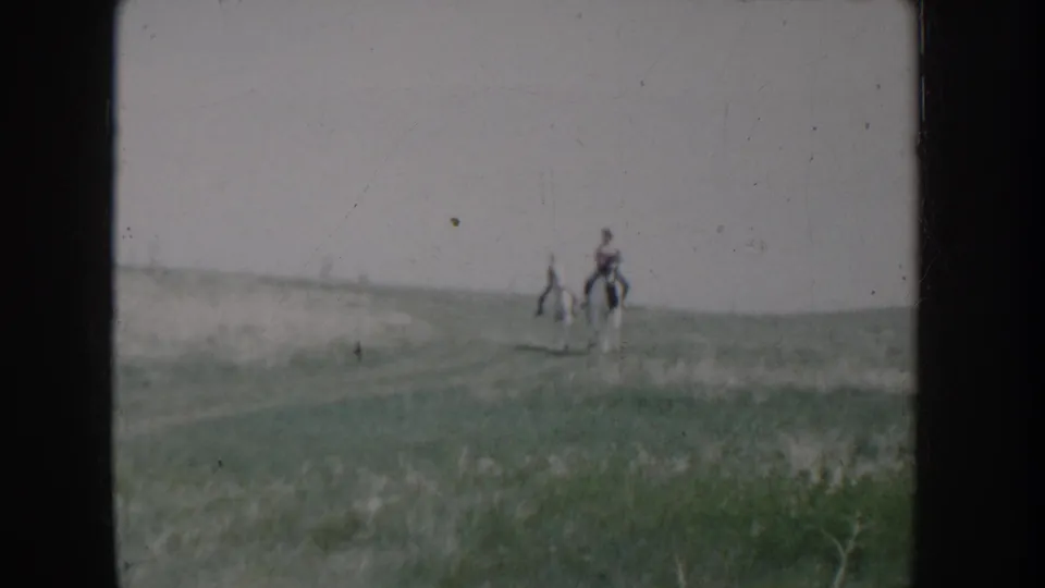 Two riders on horseback in an open grassy landscape, captured on 8mm home movie film in Idaho, circa 1955.