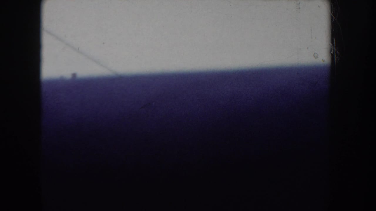 Dark 8mm frame with a large purple-toned mass dominating the foreground, small exterior scene partially visible at lower edge.