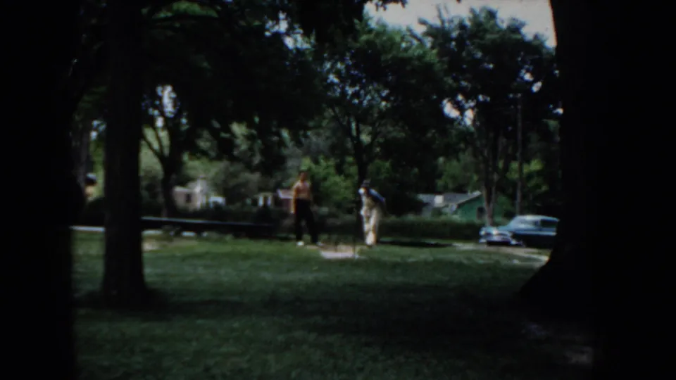Two adults in a sunlit residential yard beside a mid-1950s dark-colored automobile, large deciduous trees and homes visible in the background, Idaho, circa 1955.