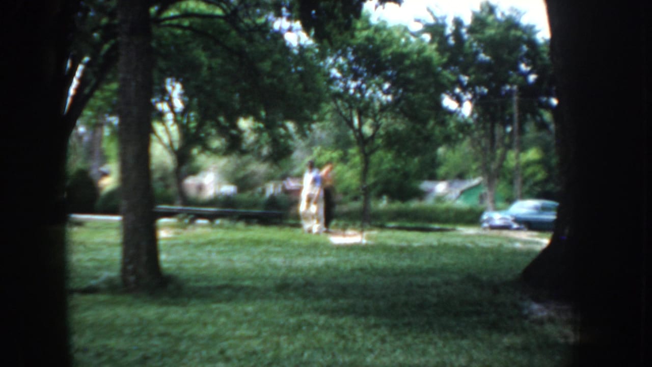 Two men standing on a green lawn between tree trunks; a period automobile is parked in the background.