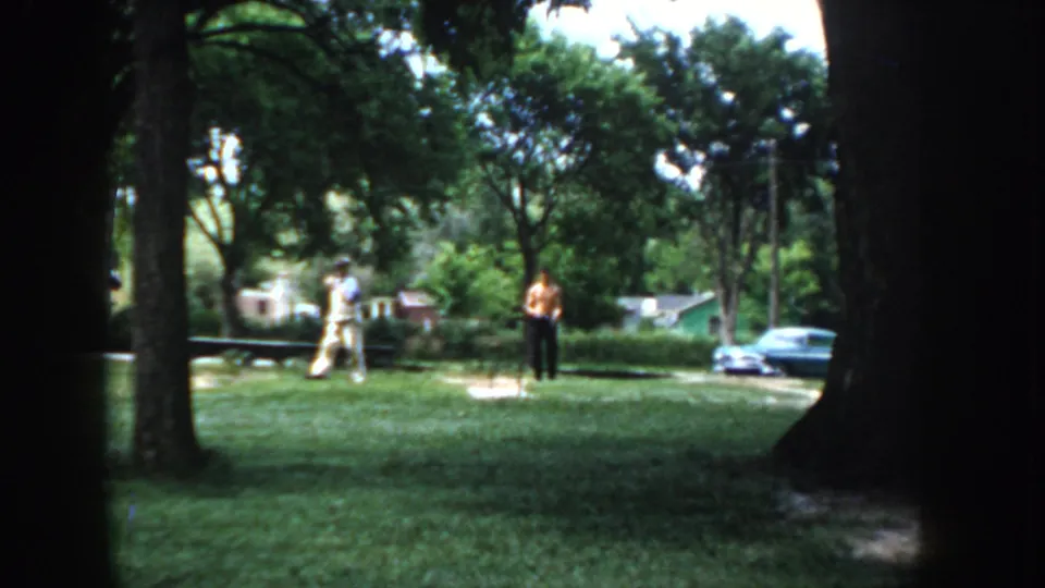 Two adult men on a summer lawn in apparent suburban Idaho, circa 1955, framed by a prominent central tree trunk, with residential structures behind on 8mm home movie film.