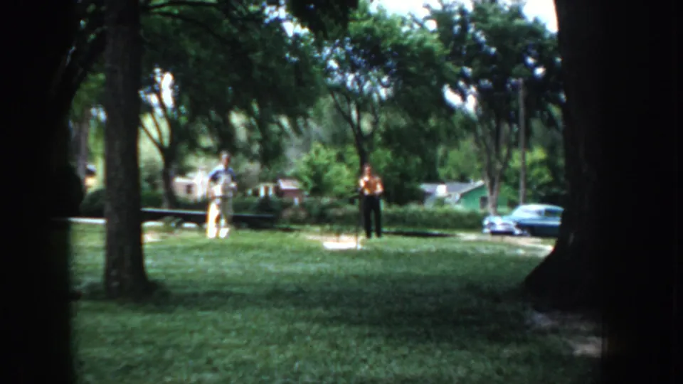 Two men on an open suburban lawn viewed through tree trunks, with period vehicles and residential structures behind; Idaho, circa 1955, on 8mm color film.