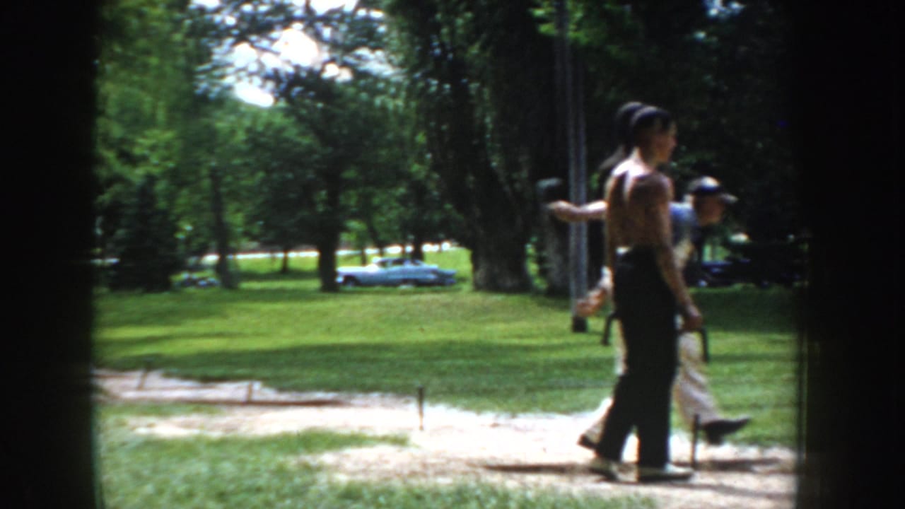 Close-mid shot of a shirtless man at ring ball stakes, circa 1955 Idaho; a shorter figure in a cap stands nearby, vintage car visible at left.