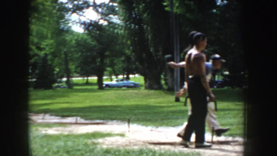 Close-mid shot of a shirtless man at ring ball stakes, circa 1955 Idaho; a shorter figure in a cap stands nearby, vintage car visible at left.