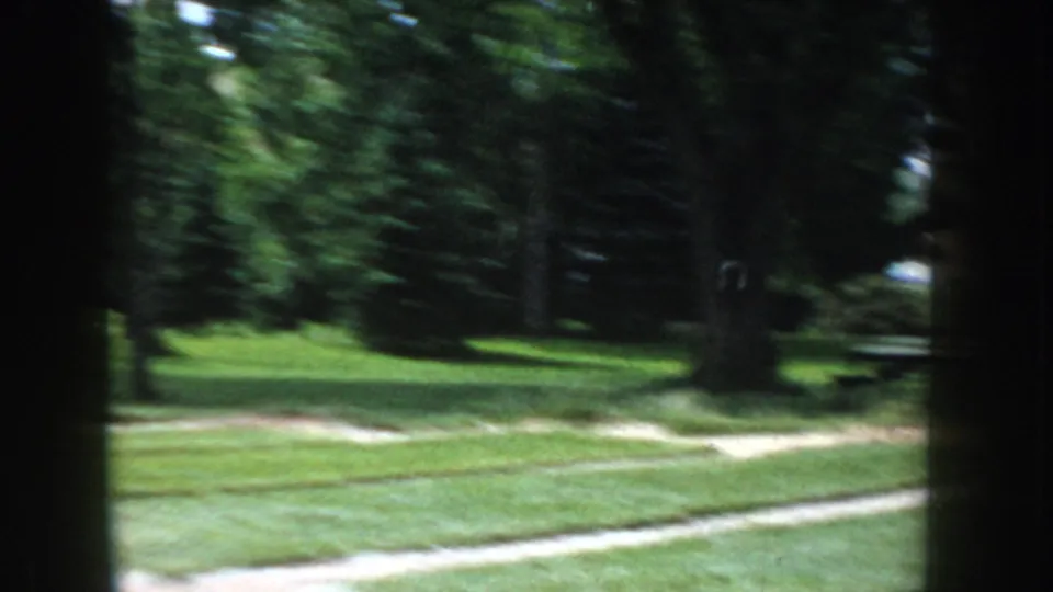 Camera pan across a tree-lined suburban park, circa 1955 Idaho; motion blur obscures detail, showing green lawn and background vehicles.