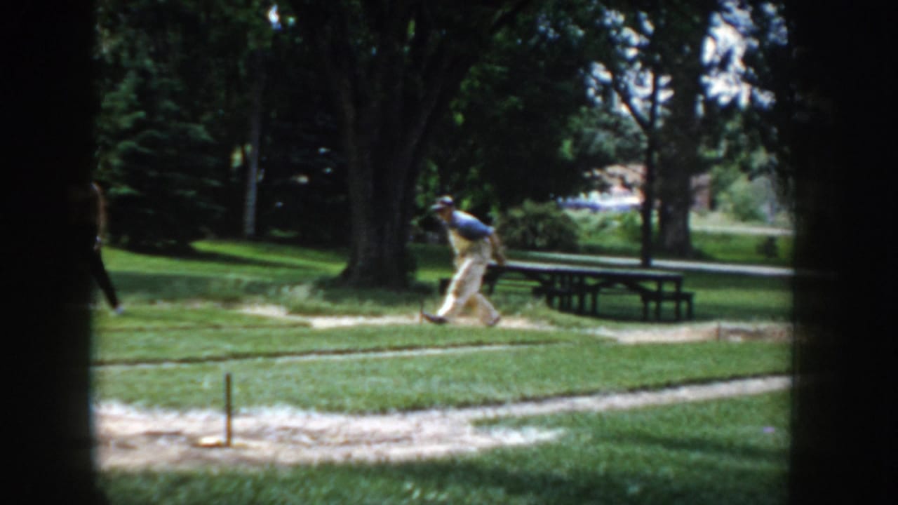 8mm home-movie frame circa 1955 showing a man in motion near a horseshoe pit in a sunny park, with wooden picnic tables and deciduous trees.