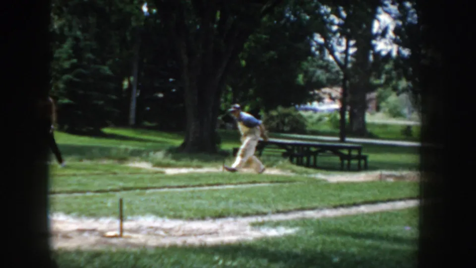 8mm home-movie frame circa 1955 showing a man in motion near a horseshoe pit in a sunny park, with wooden picnic tables and deciduous trees.