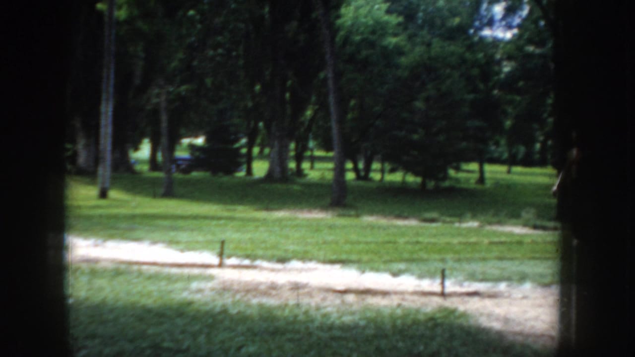 Sunny park scene showing a sandy horseshoe-pitch area with stakes, wooden picnic tables, and mature deciduous trees.