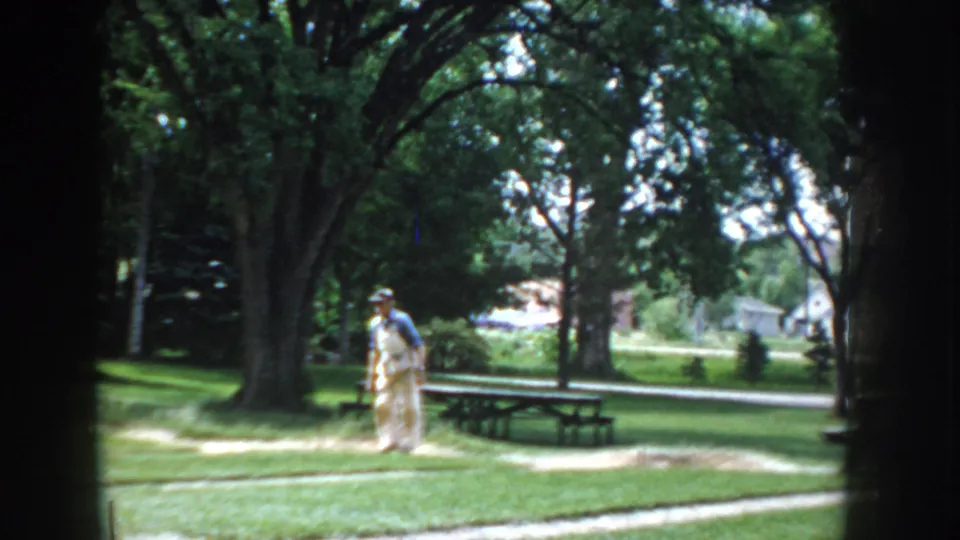 8mm home-movie frame circa 1955 showing a man in light clothing and cap standing at a horseshoe pitch in a park, picnic tables and trees visible.
