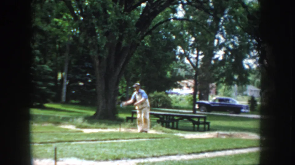 8mm home-movie frame circa 1955 showing a man in a throwing stance at a horseshoe pitch in a park; a dark period sedan and picnic tables are in the background.