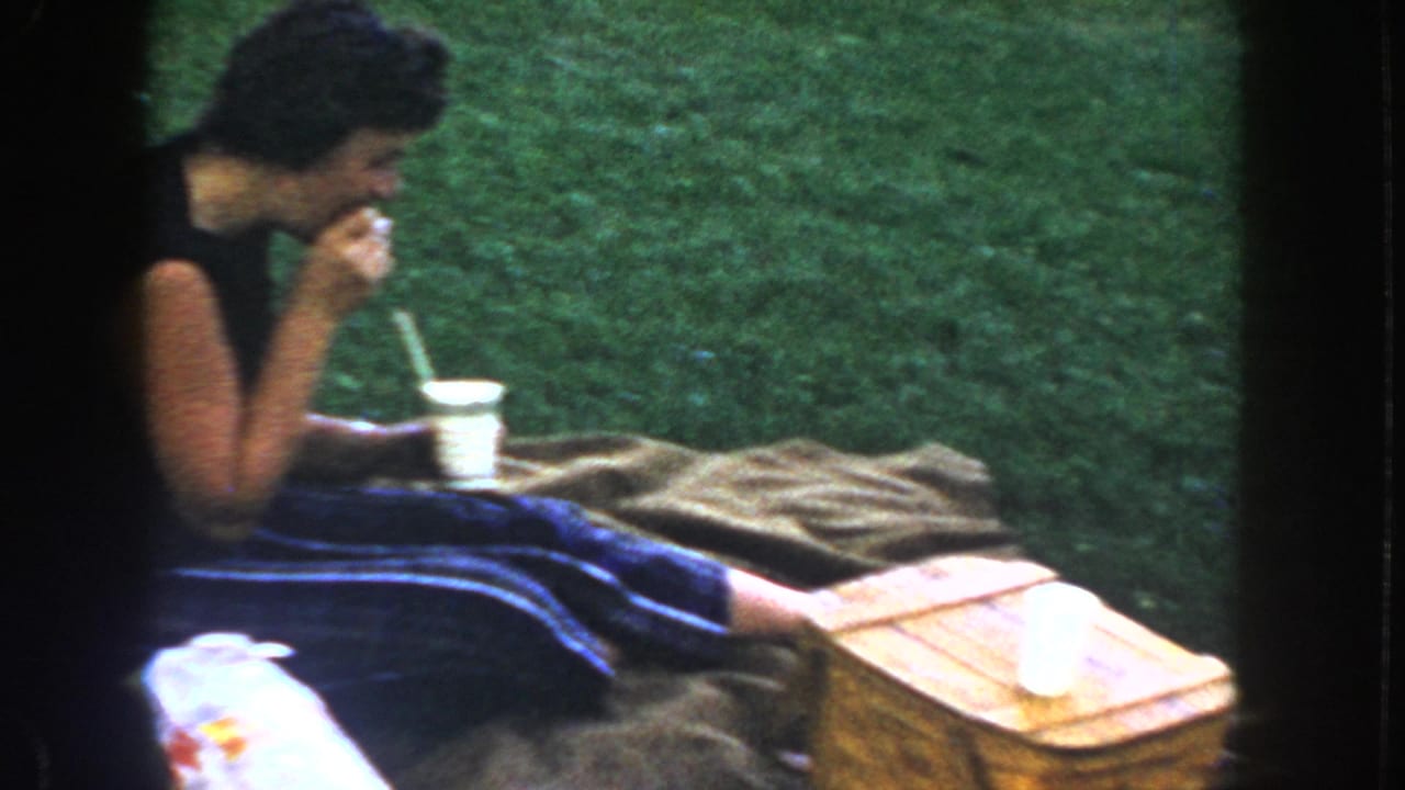 Woman in dark sleeveless dress drinks from a paper cup at a family picnic on a brown blanket on green grass, likely Idaho, circa 1955, 8mm color.