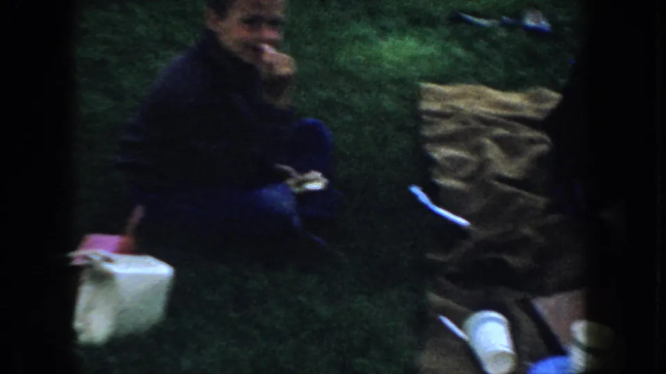 Older adult eating during a family picnic on green grass with a white box and paper cup beside them, likely Idaho, circa 1955, 8mm color footage.