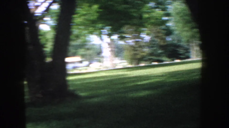 Motion-blurred 8mm frame circa 1955 Idaho: camera pans across a tree-lined suburban lawn, with light-toned vehicles or structures in a bright, partially overexposed background.