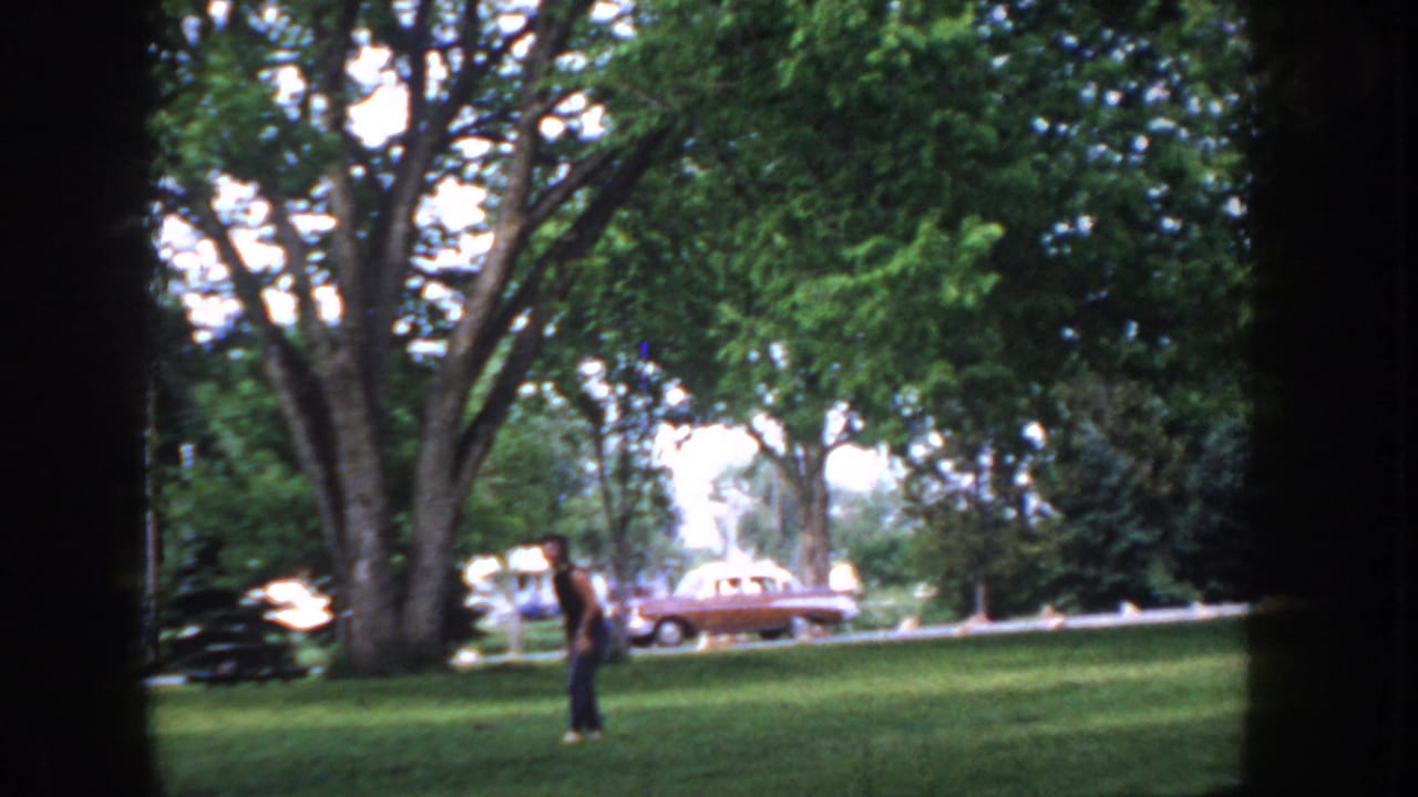 Man stands in park; a period sedan is visible further right on the road under leafy trees with open sky beyond.