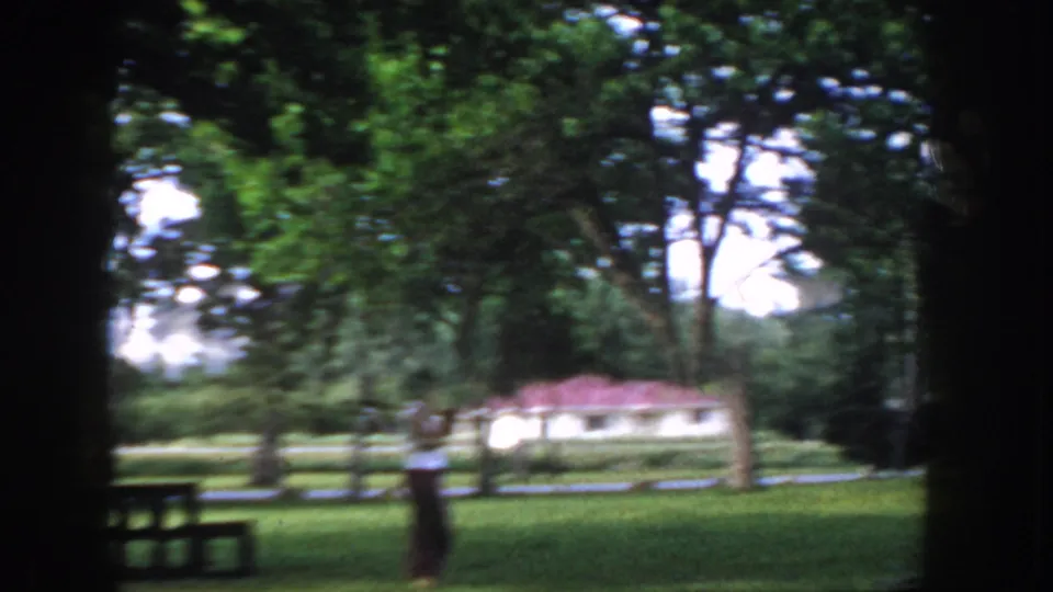 Camera has panned to show a wider park lawn with a figure in motion; a low-profile building with a reddish roof is visible in the background, with a park bench at lower left and motion blur indicating handheld filming.