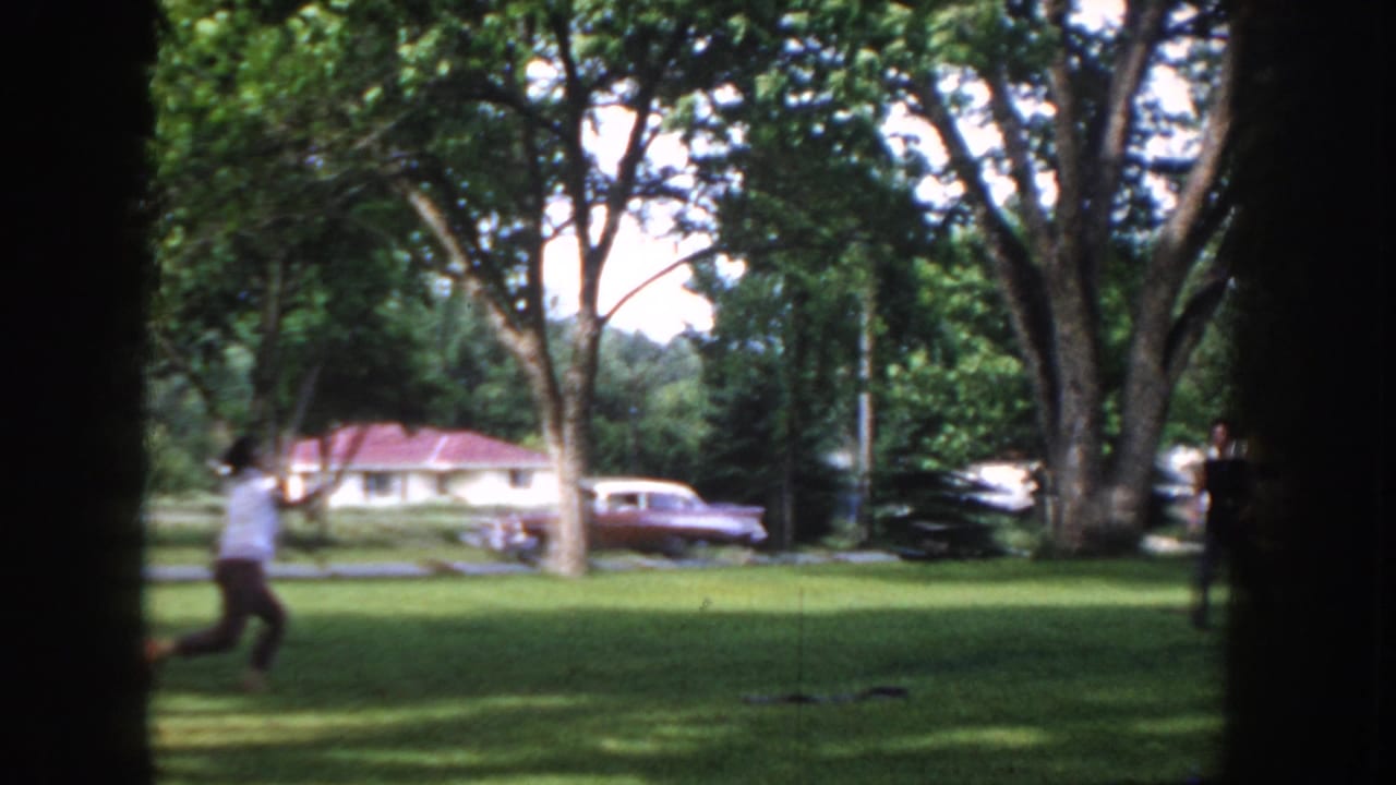 Man in 1950s casual clothing moving across a park lawn in Idaho USA, circa 1955; a period car and residential house visible behind him.