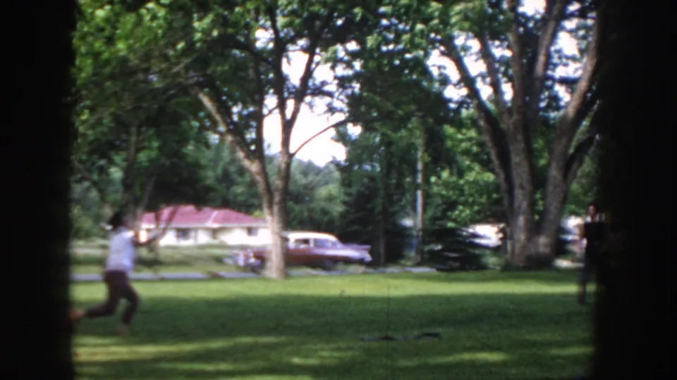 Man in 1950s casual clothing moving across a park lawn in Idaho USA, circa 1955; a period car and residential house visible behind him.