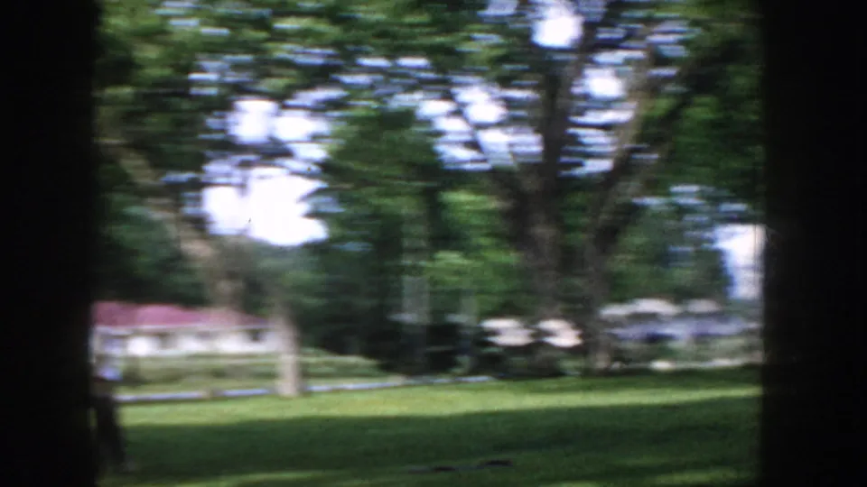 Camera pan across park trees and lawn, circa 1955 Idaho; motion blur from the sweep and a red-roofed residential building at frame left.