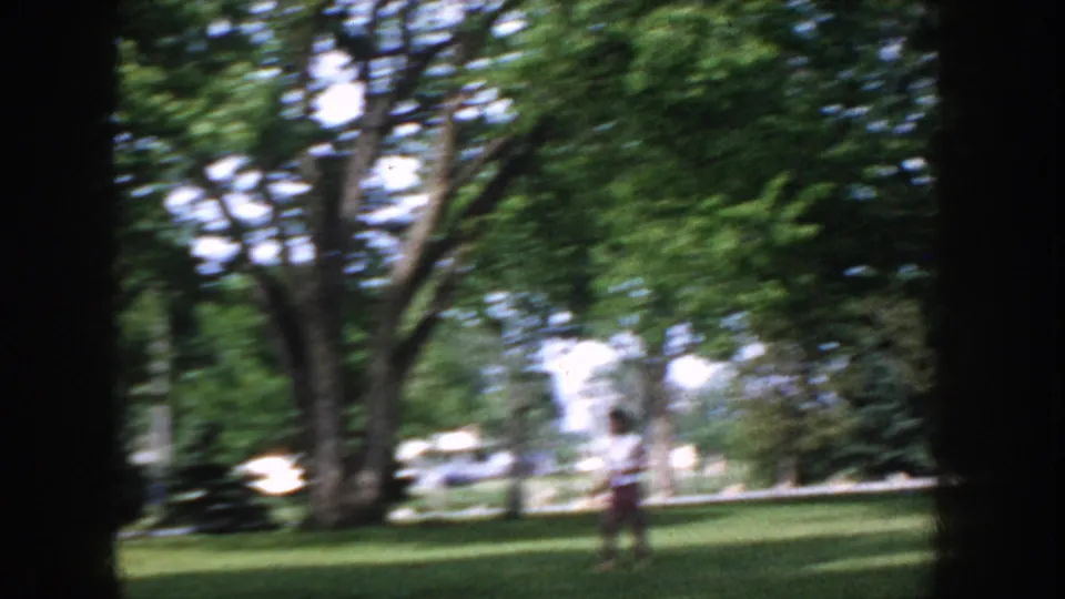 Figure in mid-century casual clothing on a park lawn, Idaho USA circa 1955; large deciduous trees and open green grass surround the figure.