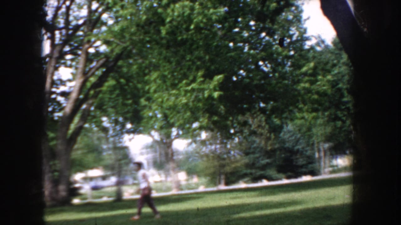 Adult pedestrian in casual clothing crosses a sun-dappled park lawn in Idaho, USA, circa 1955, with mature deciduous trees in the background.