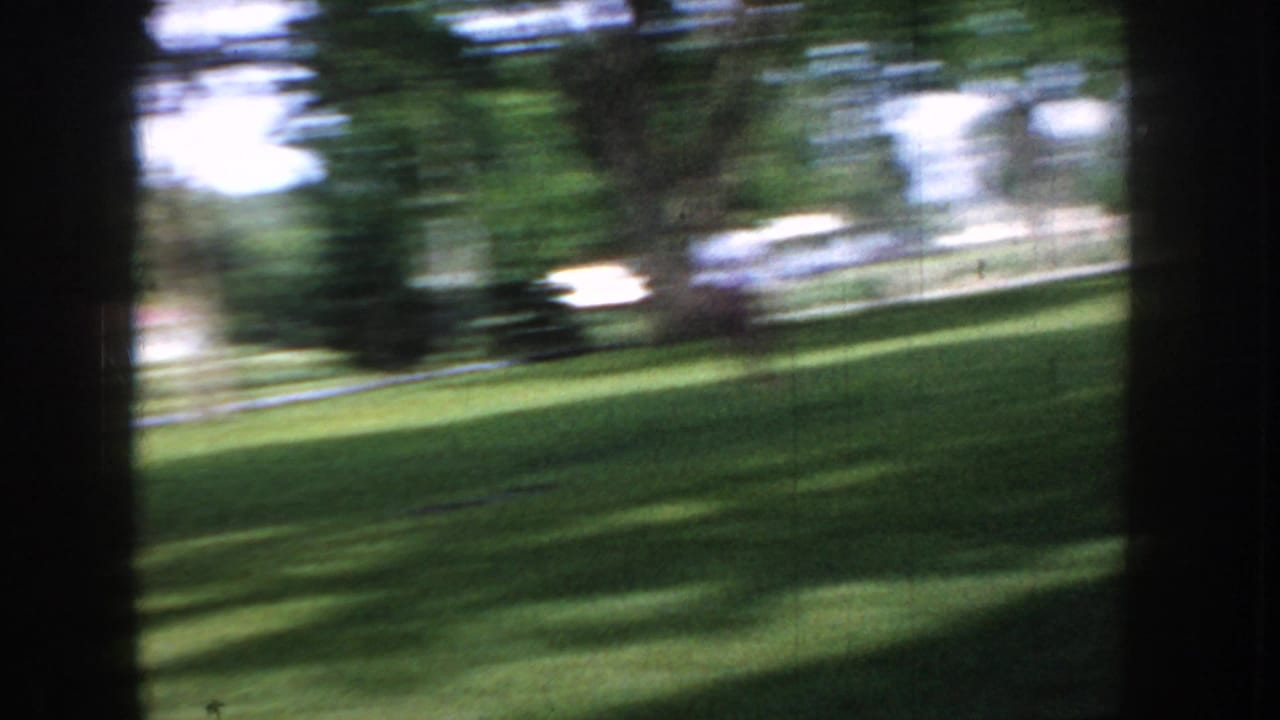 Panning shot across a sunlit park in Idaho, c. 1955, capturing a light-colored classic American car rendered in motion blur.