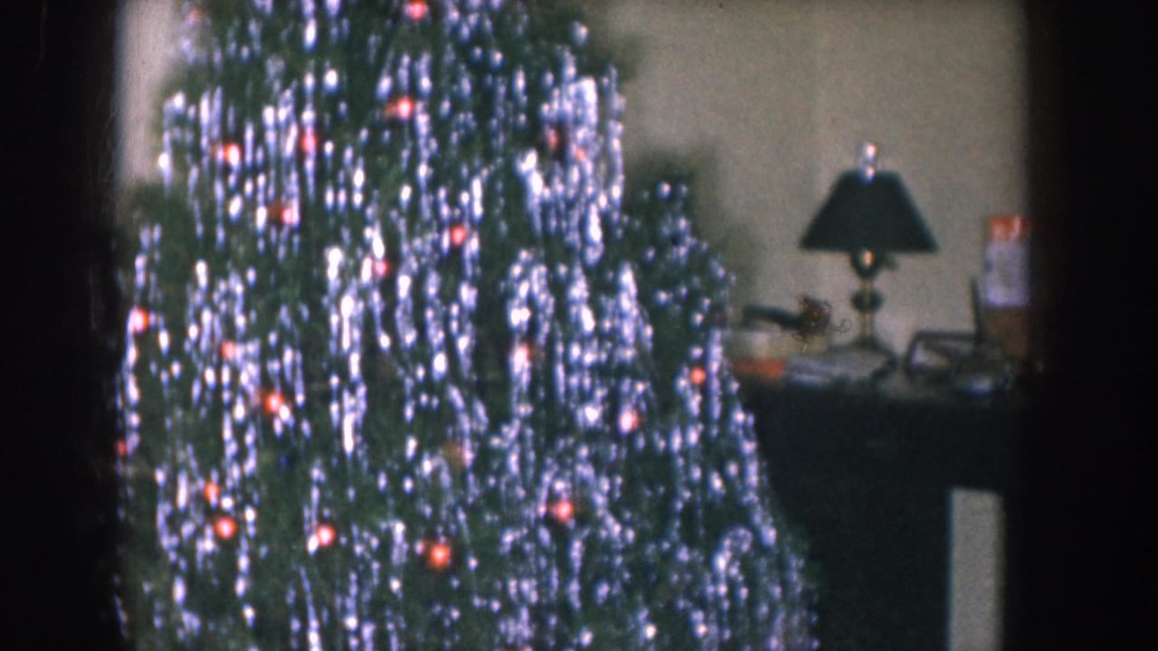 Christmas tree draped with silver tinsel and red ornaments fills the frame; a black table lamp visible in the background.