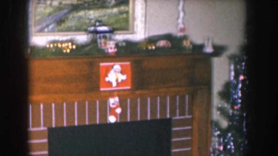 8mm home movie circa 1955 Nebraska; fireplace mantel with pine garland, framed landscape painting, Santa Claus figure, and a Christmas stocking; tinsel tree at frame left.