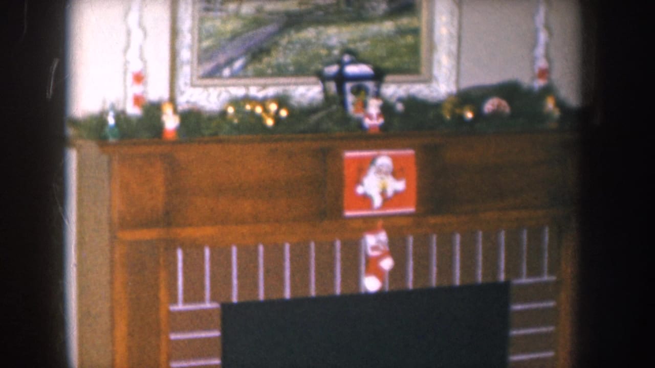 1955 Nebraska home movie frame showing a Christmas-decorated wood-panel fireplace mantel with garland, string lights, a lantern figurine, and a red stocking hung at the firebox.