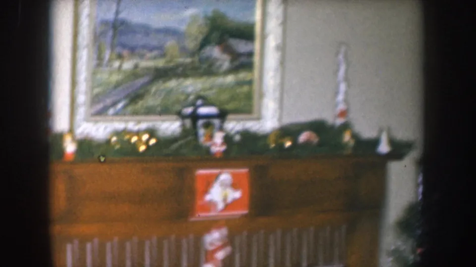 1955 Nebraska home movie frame showing a Christmas-decorated mantel with garland, lantern figurine, white cone ornaments, and a framed oil landscape painting above.