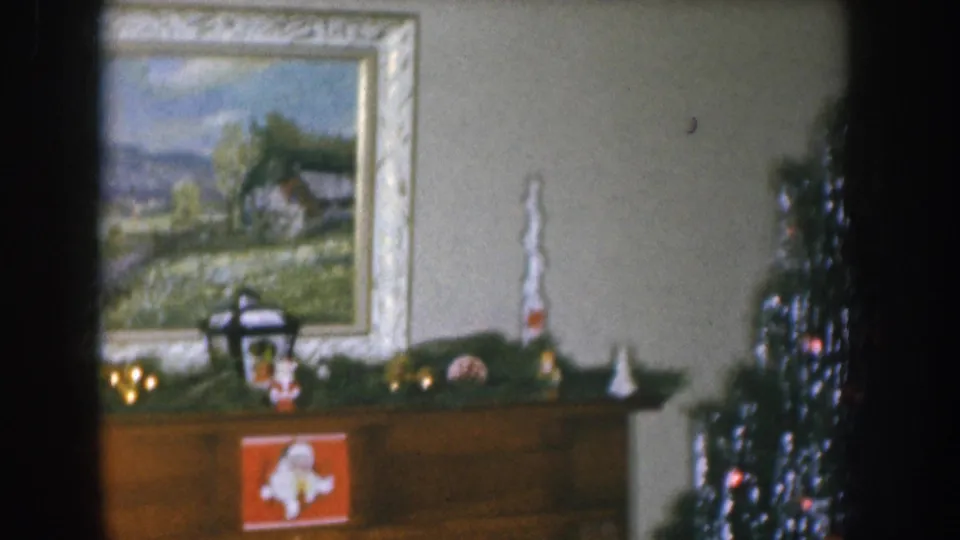 1955 Nebraska home movie frame showing a framed countryside oil painting above a garland-draped mantel, with a decorated Christmas tree partially visible at right.