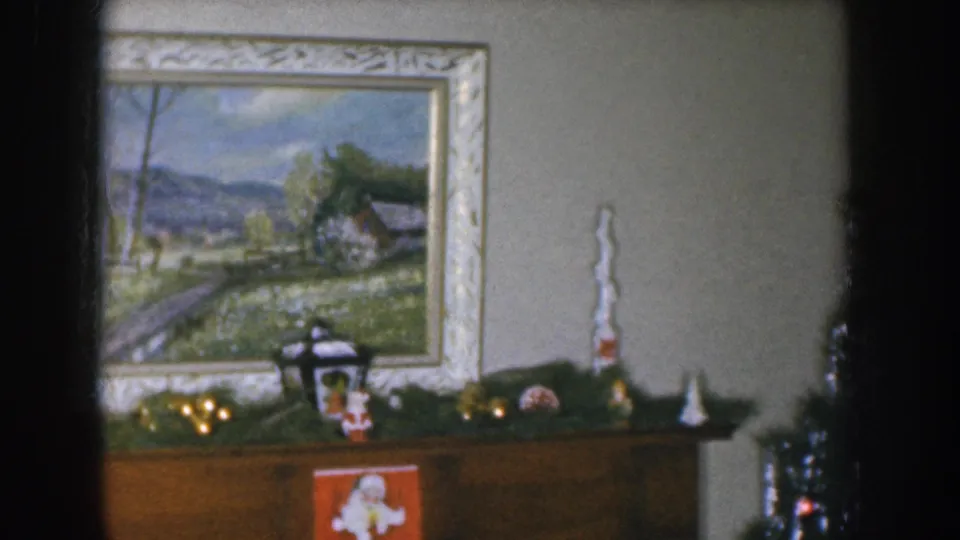 1955 Nebraska home movie frame showing a decorated Christmas tree with ornaments beside the garland-draped mantel, framed landscape painting on wall at left.