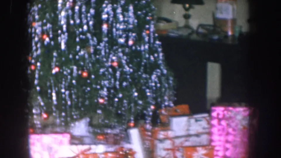 Mid-shot of Christmas tree laden with silver icicle tinsel and orange bulb lights; wrapped gifts in pink patterned, white, and other colored paper sit beneath — Nebraska, circa 1955