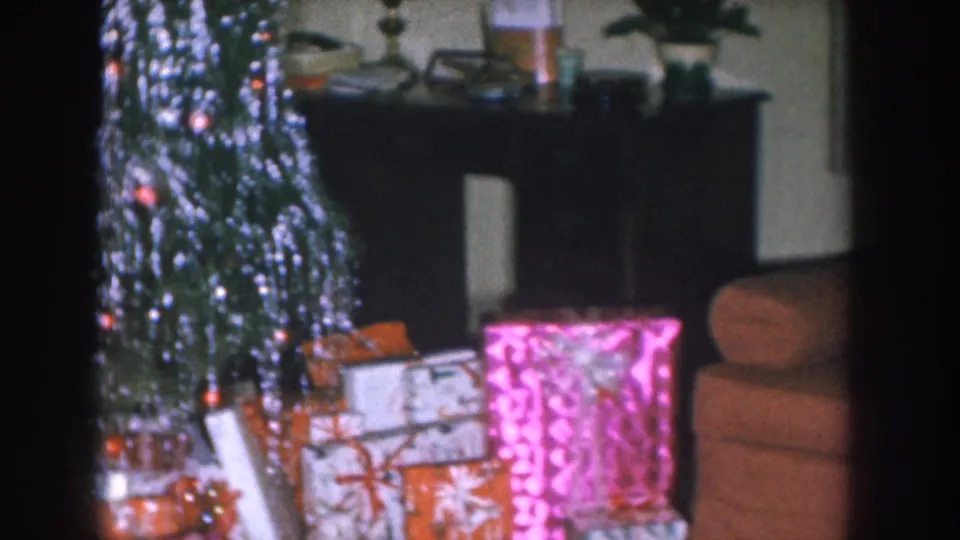 Panned view of Christmas tree with silver tinsel and orange lights; pink patterned and orange wrapped gifts in foreground, dark furniture console visible behind — Nebraska, circa 1955
