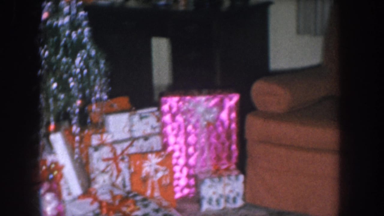 Wrapped Christmas presents including a bright pink foil box stacked near an orange armchair, with a tinsel-covered tree at left.