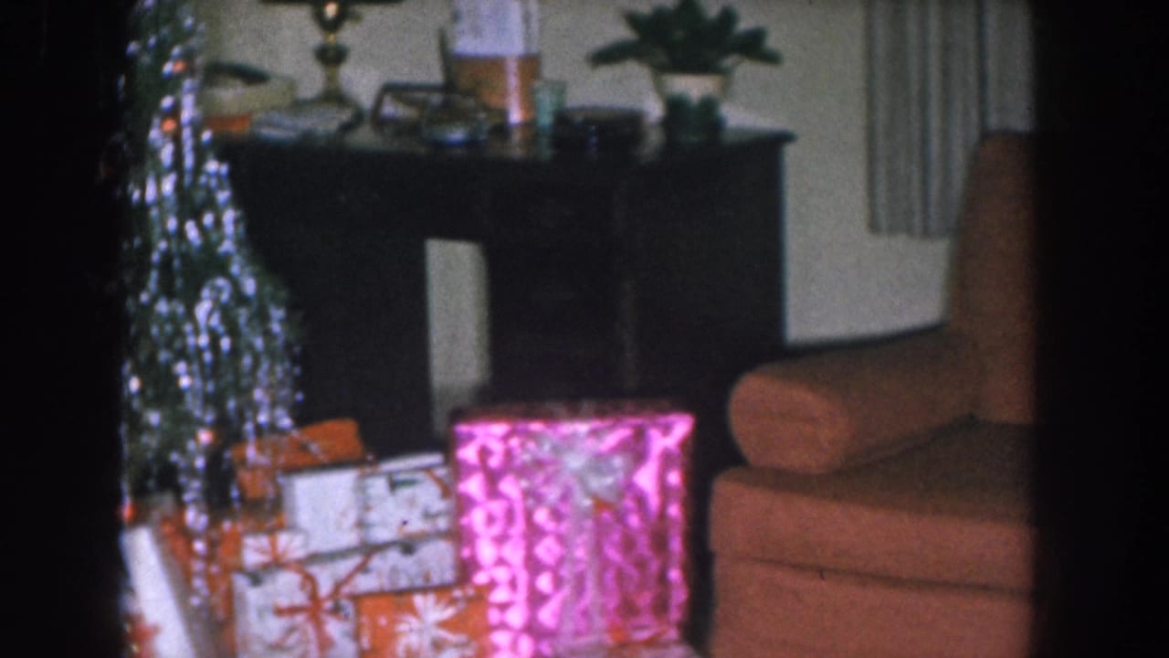 Dark wood console table visible in background above wrapped Christmas gifts, with an orange armchair and tinsel-covered tree at left.