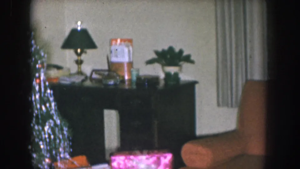 Dark mid-century console table with a black lamp, amber-coloured containers, and a small plant; tinsel tree visible at left in what appears to be a 1955 Nebraska home.