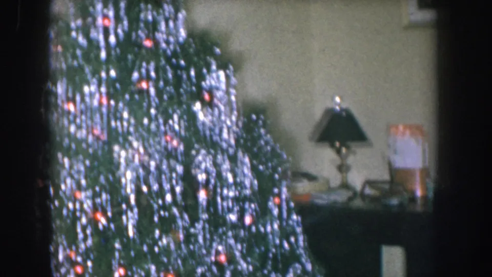 A Christmas tree covered in silver tinsel with red and orange lights glowing through, with a dark console table and lamp visible in the background of a 1950s Nebraska living room.