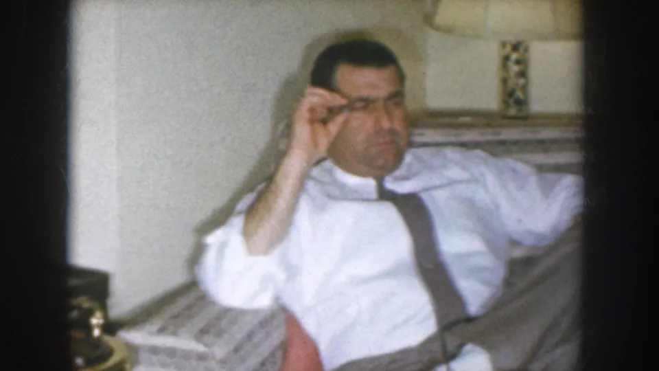Middle-aged man wearing white shirt and dark suspenders adjusting his eyeglasses while seated in a living room chair, Nebraska home movie circa 1955.
