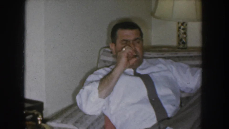 The same man raises what appears to be a cigarette to his lips while relaxing in a mid-century American living room, Nebraska circa 1955.