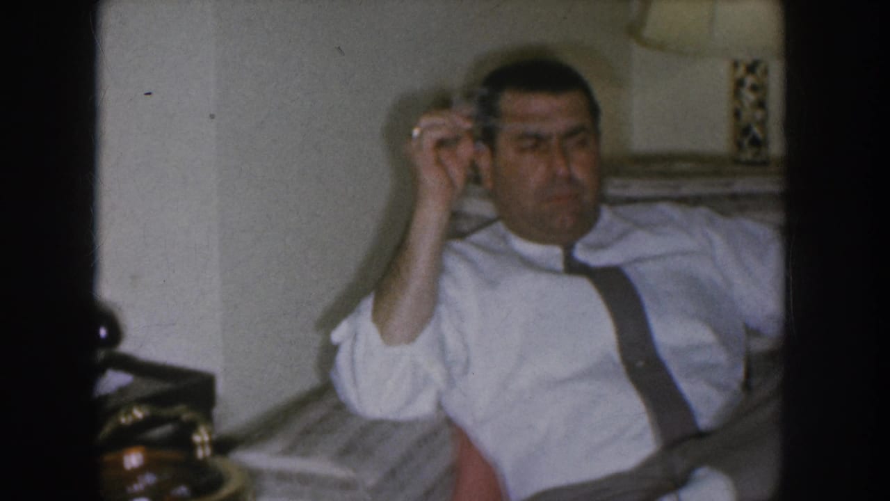 The man raises a cigarette near his head in a candid 1955 Nebraska home-movie moment; lamp with checkered shade and a dark side table visible behind him.