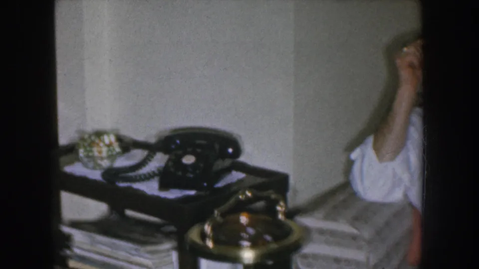 Pan shot revealing a black rotary telephone and brass ashtray on a dark wooden side table in a mid-1950s Nebraska living room, with magazines stacked beneath.