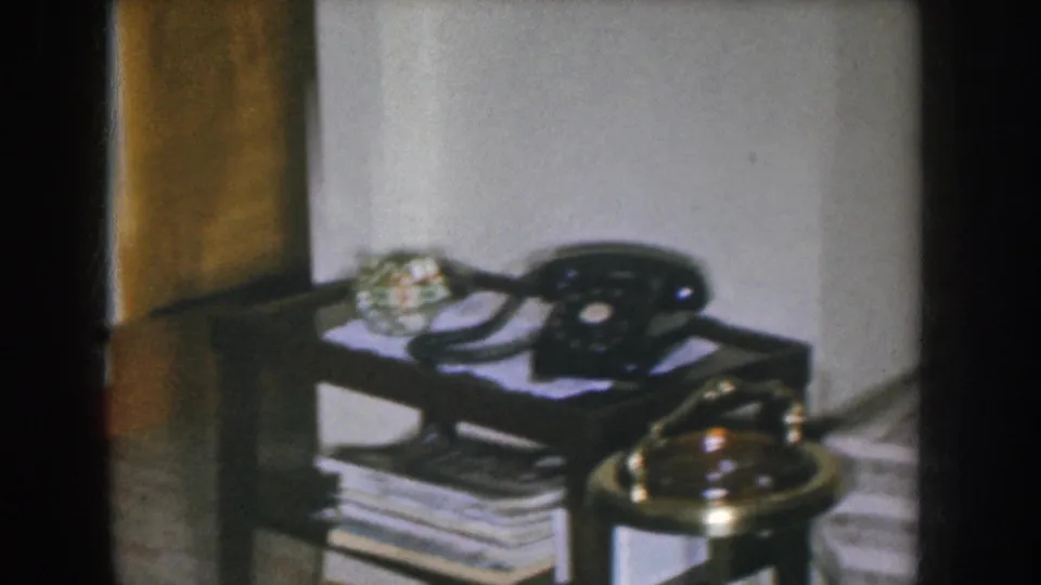 End-of-clip close shot of a black rotary telephone and brass ashtray on a magazine-stacked dark side table in a 1955 Nebraska interior; warm golden curtain drape at left.