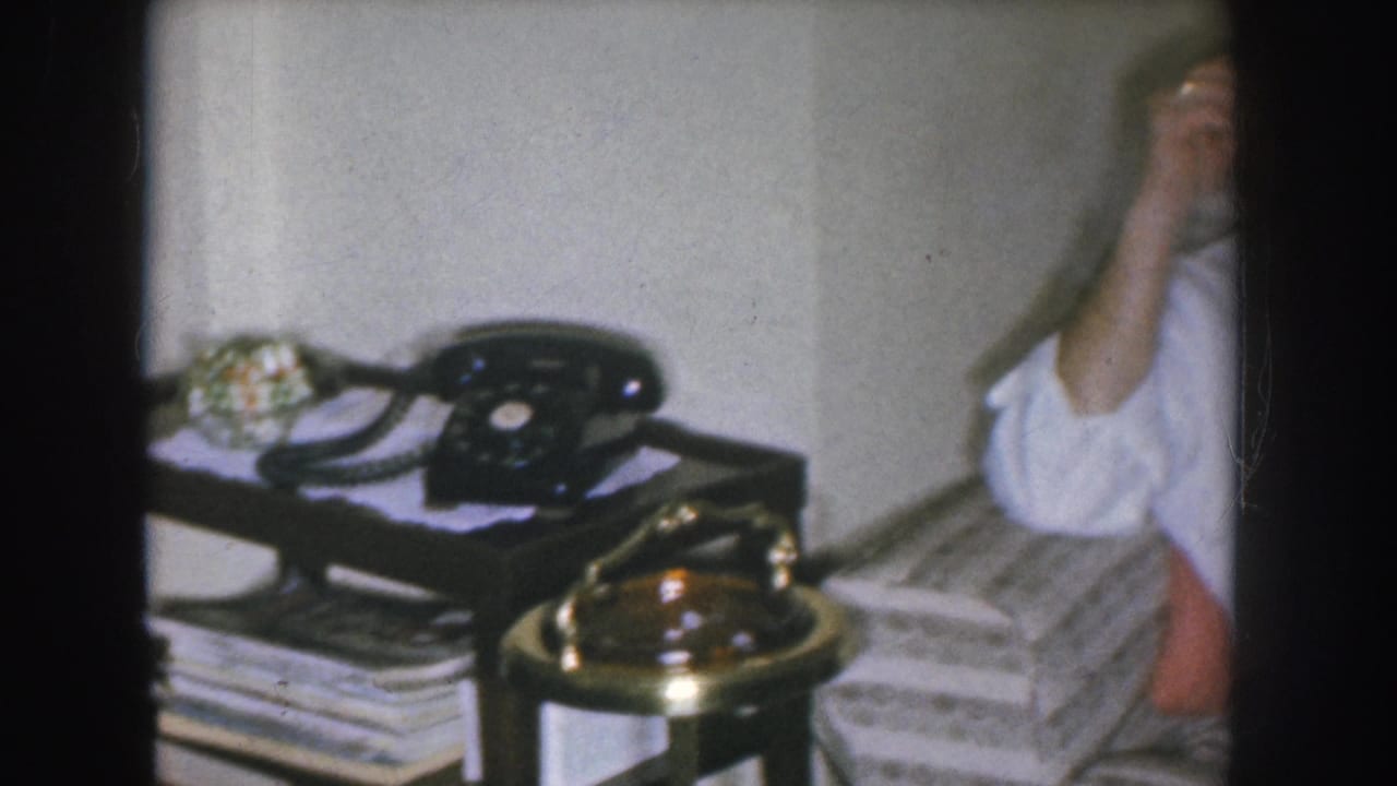 Close view of a black rotary telephone on a side table with a brass base and stacked magazines beneath, from a 1955 Nebraska 8mm home movie.