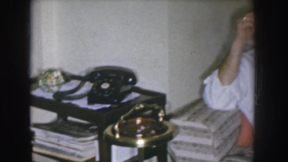 Close view of a black rotary telephone on a side table with a brass base and stacked magazines beneath, from a 1955 Nebraska 8mm home movie.