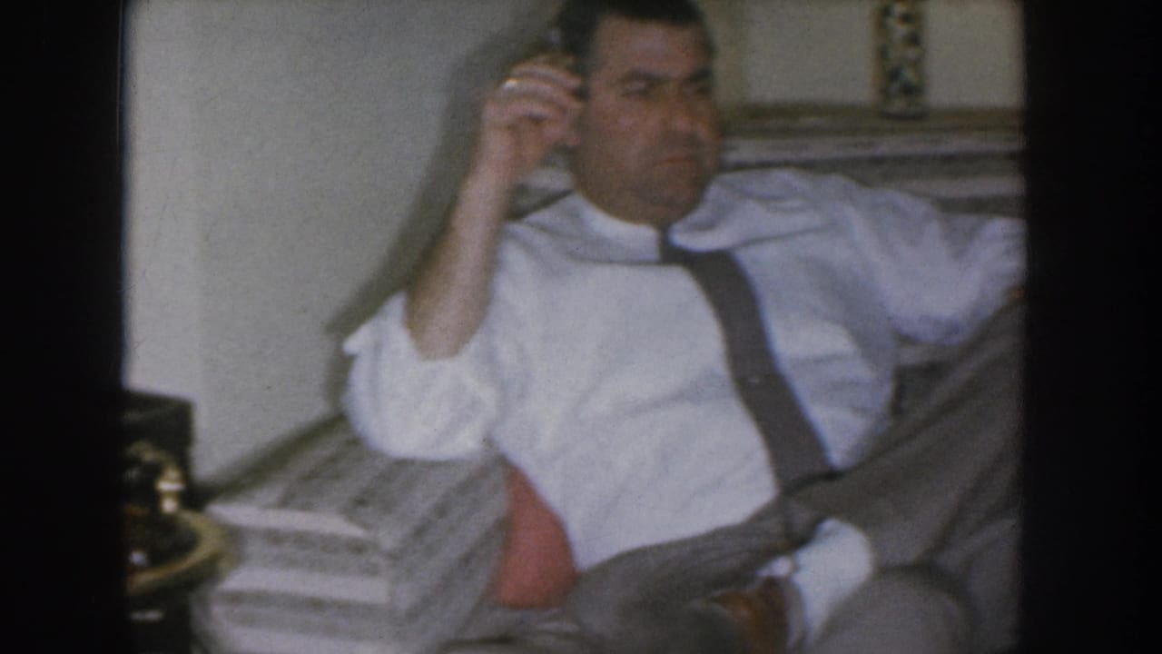 Adult man in a white dress shirt with dark suspenders holds a rotary phone receiver to his ear while seated on a sofa; Nebraska home movie circa 1955.