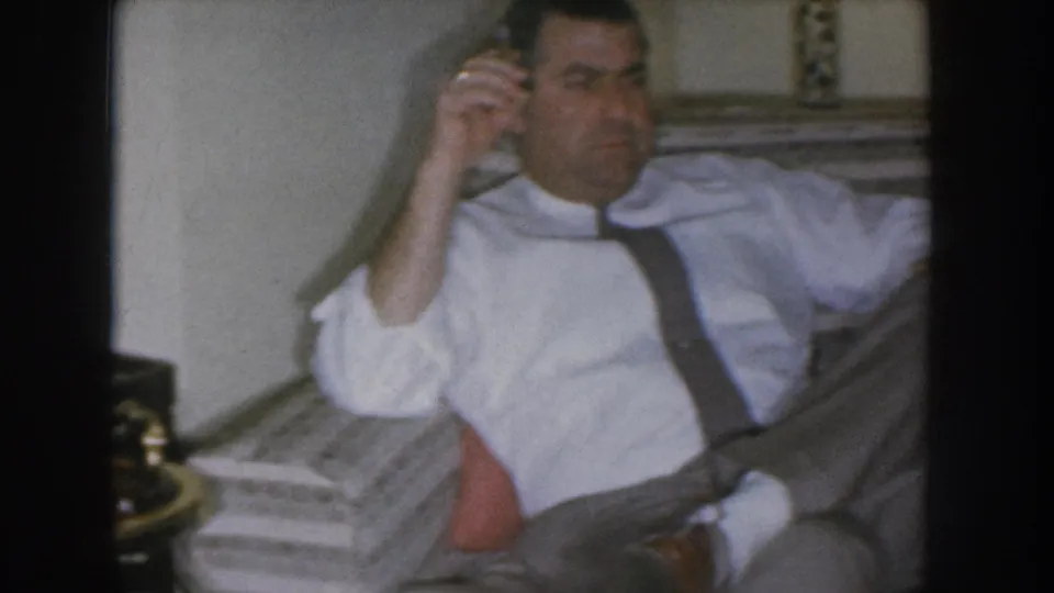Adult man in a white dress shirt with dark suspenders holds a rotary phone receiver to his ear while seated on a sofa; Nebraska home movie circa 1955.