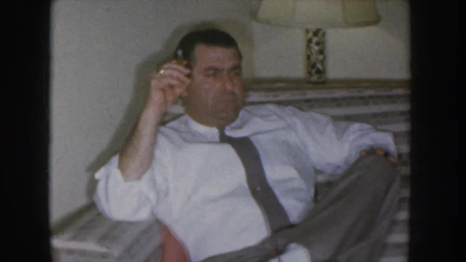 Medium-close shot of a man in a white dress shirt with dark suspenders, telephone receiver to his ear, decorative table lamp in background; Nebraska home movie circa 1955.
